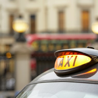 Picture shows a yellow light up sign with word 'TAXI' glowing in black on a black taxi - red bus in the background
